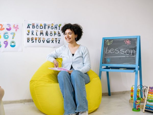 Professora sorrindo, sentada em uma sala de aula, segura um livro enquanto ensina crianças da Educação Infantil.