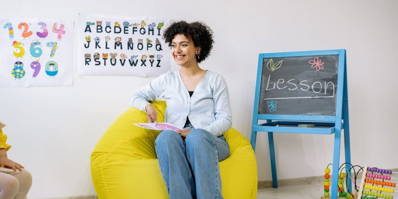Professora sorrindo, sentada em uma sala de aula, segura um livro enquanto ensina crianças da Educação Infantil.