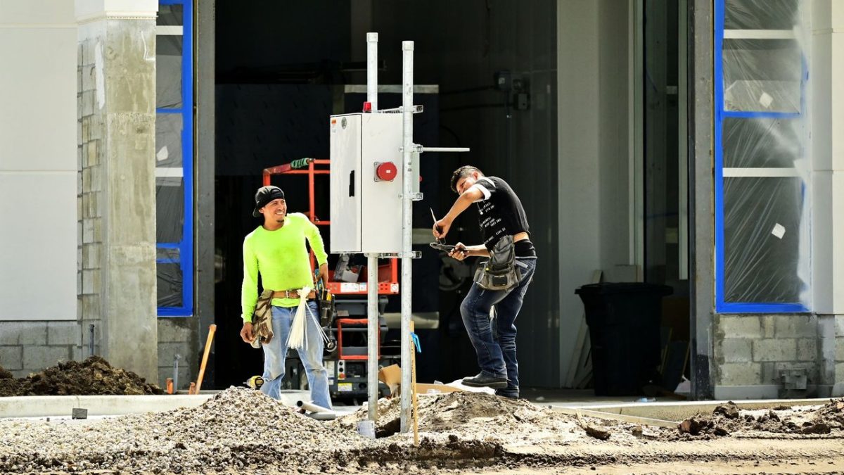 Dois trabalhadores da construção, usando equipamentos de proteção, instalam um painel elétrico em um canteiro de obras.