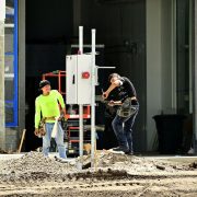 Dois trabalhadores da construção, usando equipamentos de proteção, instalam um painel elétrico em um canteiro de obras.