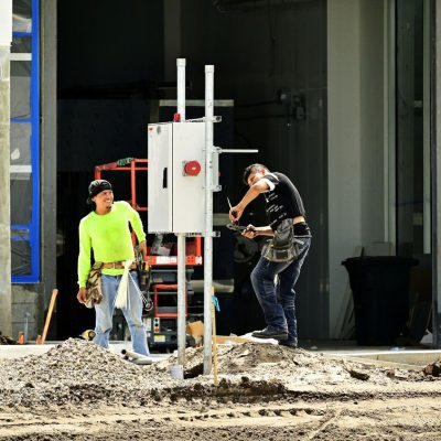 Dois trabalhadores da construção, usando equipamentos de proteção, instalam um painel elétrico em um canteiro de obras.