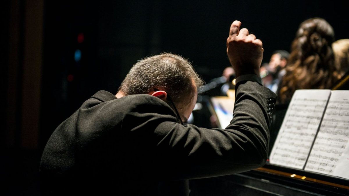 Maestro conduz uma orquestra sinfônica durante apresentação ao vivo, com partituras visíveis.
