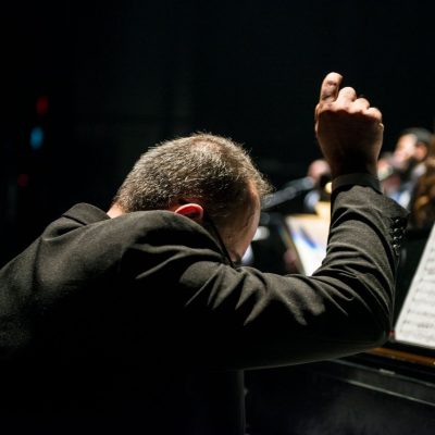 Maestro conduz uma orquestra sinfônica durante apresentação ao vivo, com partituras visíveis.