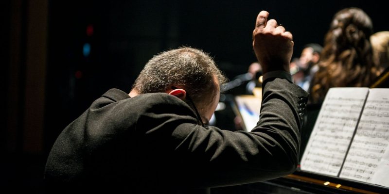 Maestro conduz uma orquestra sinfônica durante apresentação ao vivo, com partituras visíveis.
