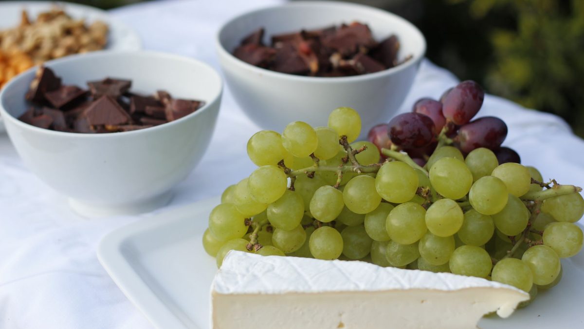 Tábua com queijos, uvas, chocolate e castanhas sobre uma mesa branca iluminada.