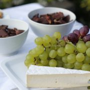 Tábua com queijos, uvas, chocolate e castanhas sobre uma mesa branca iluminada.