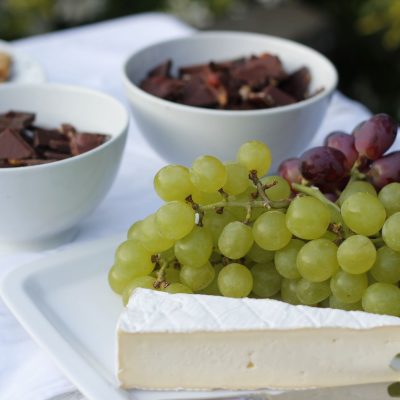 Tábua com queijos, uvas, chocolate e castanhas sobre uma mesa branca iluminada.