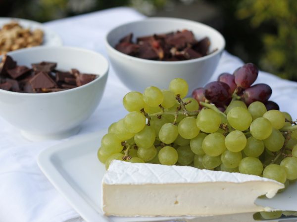 Tábua com queijos, uvas, chocolate e castanhas sobre uma mesa branca iluminada.