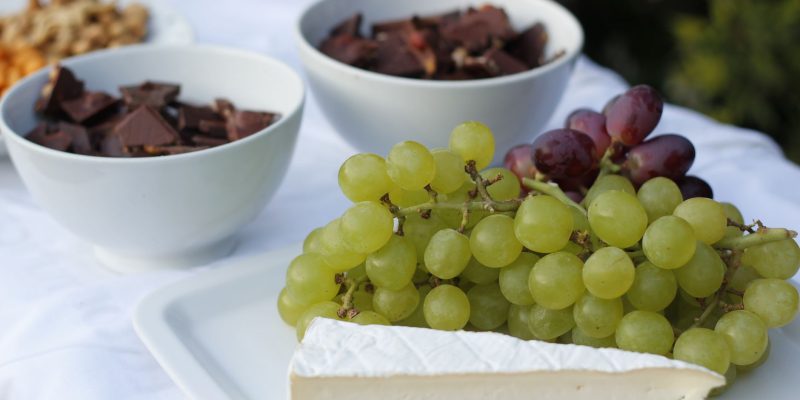 Tábua com queijos, uvas, chocolate e castanhas sobre uma mesa branca iluminada.